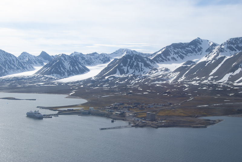 Kystsamfunn i arktis omgitt av snødekte fjell, med en cruiseskip ved kai, spredte bygninger langs strandlinjen, og rolig vann under delvis skyet himmel.